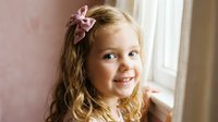 Happy blonde toddler girl with curly hair and pink bow smiling by window in soft natural light for family portraits and childhood memories.