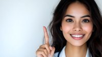 Young Asian woman with bright smile pointing finger up against light background, expressing positive attitude and confidence. Professional portrait for business.