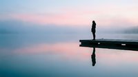 Woman standing on wooden dock at dawn, silhouetted against calm water and pastel sky. Serene lakeside scene for wellness, mindfulness, and contemplation concepts.