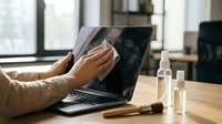 Professional woman cleaning laptop screen with microfiber cloth at modern office desk with sanitizer bottles for workplace hygiene and device maintenance.