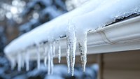 Frozen icicles hanging from white house gutter against winter sky background, showing seasonal weather conditions and potential roof maintenance needs.