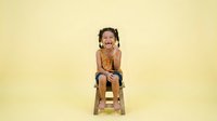 Young child with dark curly hair and medium skin tone sitting on wooden stool against yellow background, wearing mustard-colored sleeveless shirt, expressing joy and excitement for lifestyle and parenting content.