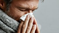 Mature Caucasian man with gray scarf blowing nose into tissue, showing signs of cold or flu against neutral gray background.