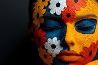 Close up portrait with yellow and blue face paint decorated with red and white floral elements, closed eyes and orange lips against dark background.