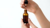 Female hands holding amber glass dropper bottle dispensing essential oil or serum against white background for skincare and wellness applications.