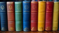 Row of vintage hardcover books with colorful spines in blue, green, red, yellow and brown shades arranged on shelf, showing classic binding details and gold embossed designs.