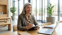 Professional mature Asian businesswoman smiling while using smartphone at modern office desk with laptop and documents in bright workspace.