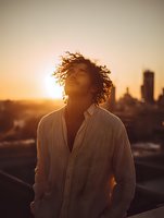 Young African American man with curly hair standing against urban sunset skyline, eyes closed in peaceful moment, wearing casual striped shirt, backlit by golden hour light.