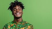 Young African American man with dreadlocks laughing joyfully while wearing tropical print shirt against green background, expressing genuine happiness and positive emotions.
