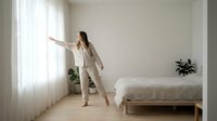 Woman in beige linen suit stretching arms in minimalist bedroom with natural light, wooden floor, potted plant, and bed for wellness and lifestyle content.