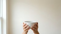 Hands holding empty white ceramic bowl against neutral beige wall with natural light, suitable for food, kitchen, dining, and lifestyle content.