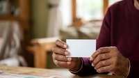 Mature man with dark skin tone wearing burgundy sweater holding blank white card in hands indoors, suitable for business communication and greeting card concepts.