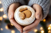 Female hands in gray sweater holding white cup with gingerbread cookies and cinnamon stick, sprinkled with cocoa powder against blurred festive lights background.