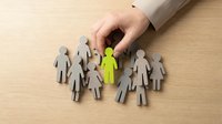 Hand selecting a green figure among gray figures on wooden surface, representing recruitment, talent selection, hiring, and human resources management concepts.