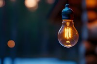 Glowing vintage filament light bulb hanging against blurred bokeh background, warm amber illumination creates atmospheric mood in evening setting.