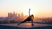 Young woman practicing yoga side plank pose on rooftop at sunrise with city skyline backdrop for fitness wellness lifestyle content.