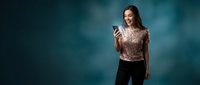 Excited young Caucasian woman in sequin top using smartphone, glowing screen, dark teal background, copy space for text.