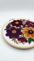 Embroidered floral hoop art featuring vibrant purple, burgundy, and golden yellow flowers with green leaves and decorative beads on white fabric in wooden frame.