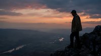 Silhouetted hiker standing on mountain peak at sunset, overlooking vast valley landscape with winding river below and dramatic sky. Adventure and outdoor exploration.