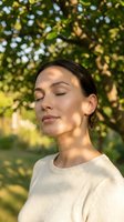 Peaceful young woman with closed eyes enjoying warm sunlight filtering through green tree leaves in natural outdoor setting for wellness and mindfulness content.