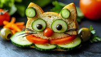 Creative cat-shaped sandwich with cucumber eyes, olive pupils, tomato nose, and cucumber smile garnish on dark slate background, fun food art for children.