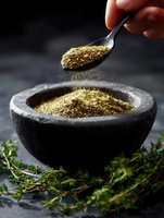 Dried aromatic herbs and spices in stone mortar with spoon sprinkling seasoning powder, fresh thyme sprigs on dark background, macro food photography.