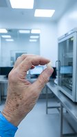 Senior male scientist holding small white pharmaceutical capsule in modern laboratory setting with medical equipment and bright lighting.