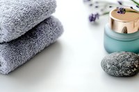 Soft gray folded towels with spa cosmetic jar and massage stone on white background, lavender flowers for wellness and relaxation concept.