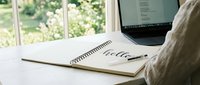 Spiral notebook with hello lettering on a wooden desk beside a laptop, cozy home workspace with bright garden window background.