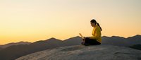 Woman in yellow jacket sitting on mountain peak with laptop at sunset, working remotely in nature with golden sky and distant hills.