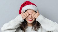 Young woman in Santa hat covering eyes with hands, wearing white sweater against gray background, smiling with red lipstick for holiday season.