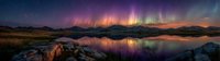 Spectacular aurora borealis panorama over calm lake at night, colorful northern lights reflecting in water with mountains and rocky shore in foreground.