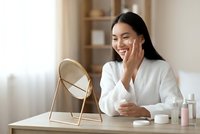 Young Asian woman applying skincare cream while looking in mirror at home beauty routine for healthy glowing skin care and wellness lifestyle.