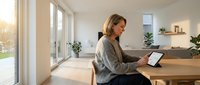 Young blonde woman using tablet computer at wooden table in modern bright living room with large windows and natural sunlight streaming in.