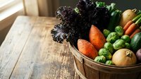 Wooden basket filled with fresh organic vegetables including dark leafy greens, carrots, peas, cucumbers and onions on rustic table for cooking and healthy eating.