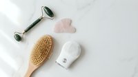 Spa wellness tools with jade roller, gua sha stone, dry brush and moisturizer on white marble background for skincare routine and self-care rituals.