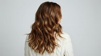 Young woman with long wavy brown hair from behind wearing white sweater against gray background for hair care and beauty concepts.