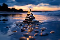Balanced stone pyramid on beach at sunset with dramatic orange sky reflecting in wet sand and shallow water, creating zen-like atmosphere and tranquility.