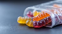 Colorful gummy candy worms spilling from clear plastic package on dark background, macro shot showing sugar coating and translucent texture. Suitable for food marketing.