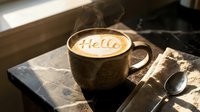 Steaming latte art with Hello written on frothy milk in a rustic ceramic mug on a marble table in warm morning light.