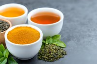 Fresh green tea leaves, brewed tea, and turmeric powder in white ceramic bowls on dark background with mint leaves, arranged for wellness and cooking.