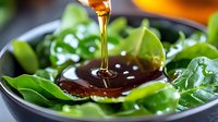 Honey drizzling onto fresh green salad leaves in dark ceramic bowl, close-up view with selective focus showing golden syrup stream and droplets.