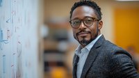 African American businessman wearing glasses and grey suit jacket smiling confidently in modern office setting with blurred background, professional corporate portrait.