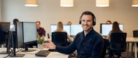 Smiling male customer service representative with headset working at computer in modern office environment with colleagues in background.