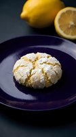 Homemade lemon crinkle cookie dusted with powdered sugar on dark blue plate with fresh lemons in background for baking and dessert recipes.