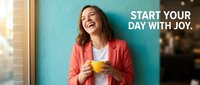 Happy businesswoman in red blazer holding yellow coffee cup laughing joyfully against turquoise wall in modern office space for morning motivation campaigns.