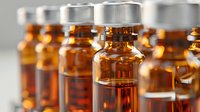 Rows of amber glass medical vials with aluminum caps containing liquid medicine or vaccine, shallow depth of field focus on front bottles in laboratory setting.