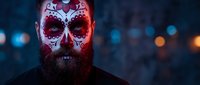 Bearded man with traditional sugar skull face paint featuring red and white decorative patterns for Day of the Dead celebration against blurred city lights.
