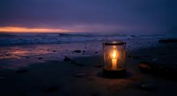 Glowing lantern on rocky beach at twilight with dramatic purple and orange sunset sky reflecting on calm ocean waves creating peaceful atmospheric mood.