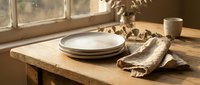 Rustic wooden table with stacked white plates, linen napkin, dried botanicals and ceramic mug near window for dining and home decor styling.